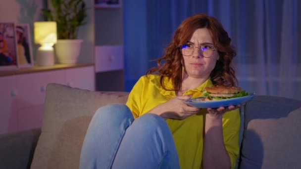 Vrouw met bril is kijken tv-show en het eten van junk food hamburger terwijl ontspannen in de woonkamer in de avond, quarantaine — Stockvideo