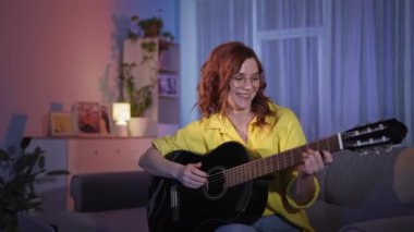 Gözlüklü kadın internette gitar çalmak için dizüstü bilgisayar kullanmayı akşamları evde oturarak öğreniyor.