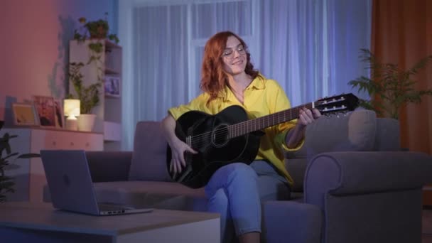 Mujer tocando una guitarra instrumental estudiando música y notas en el portátil mientras se sienta en el sofá en casa por las noches en auto-aislamiento durante la cuarentena — Vídeo de stock