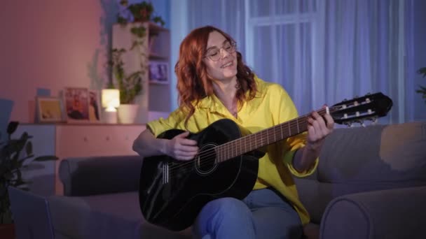 Dziewczyna śpiewa piosenki i gra na gitarze instrumentalnej, studiuje nuty i muzykę online za pomocą laptopa podczas siedzenia na kanapie w domu późnym wieczorem — Wideo stockowe