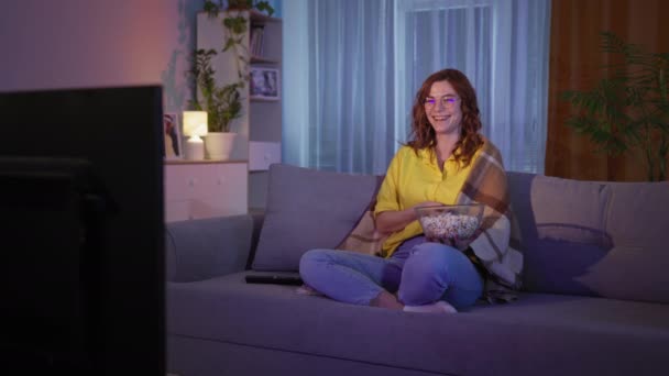 Menina alegre comer pipocas ri e se diverte assistindo TV enquanto sentado no sofá na sala de estar no final da noite — Vídeo de Stock