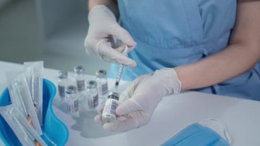 Doktor masaya yakın oturuyor, elinde koruyucu eldivenlerle şırıngayla aşıyı camdan şişeyle dolduruyor.