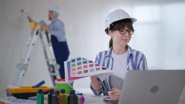Mädchen mit Helm und Brille arbeitet am Computer und wählt neue Tapeten für Wände Hintergrund des männlichen Malers im Zimmer — Stockvideo