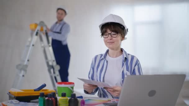 Femme avec palette de couleurs choisir la couleur de la peinture tout en étant assis à l'ordinateur et pointe son doigt vers le mur tout en parlant à l'homme faisant des réparations à l'intérieur — Video