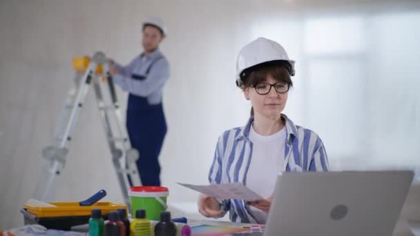 Femme en casque dur et lunettes avec palette de couleurs choisit la nouvelle conception de la pièce tout en étant assis à l'ordinateur et pointant du doigt le mur tout en parlant au constructeur masculin à l'intérieur — Video