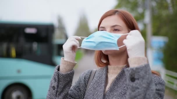 Mujer toma precauciones y se pone máscara médica y guantes para protegerse contra la infección por virus y coronavirus durante la pandemia y la distancia social telón de fondo de los autobuses urbanos — Vídeo de stock