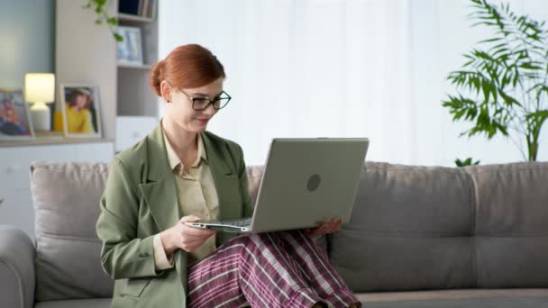 Kvinna med glasögon i jacka och pyjamas byxor arbetar online på laptop i hemmakontoret under karantän — Stockvideo