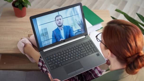 Kobieta pracownik komunikuje się za pośrednictwem wideokonferencji na laptopie z kolegami pracującymi zdalnie w domu, siedząc na kanapie w kurtce i piżamie w pokoju — Wideo stockowe