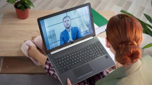 Empleada se comunica a través de videoconferencia en el ordenador portátil con sus colegas mientras está sentada en el sofá con chaqueta y pijama en la habitación — Vídeos de Stock