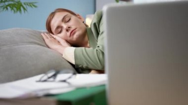 Kadın ceketli, battaniyeyle örtülmüş bir şekilde kanepenin yanında dizüstü bilgisayar ve evdeki evraklarla karantina altında yatıyor.