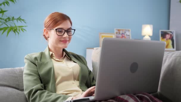 Vrouw in bril in jas en pyjama werkt online achter laptop op de bank, glimlachen en wijzen naar camera — Stockvideo