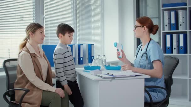 Familia en la cita con el médico, médico femenino mide la temperatura de un niño pequeño paciente con un termómetro infrarrojo sin contacto — Vídeo de stock