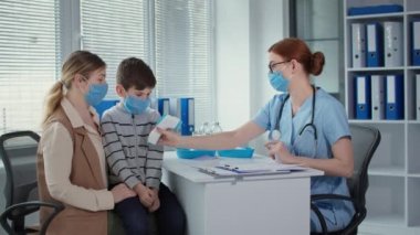 masked family at the doctor appointment, female physician measures the temperature of little boy patient with a non-contact infrared thermometer before the injection procedure