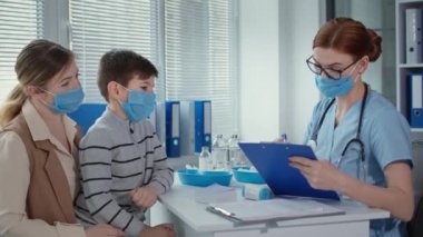 healthcare, masked doctor with clipboard measures the temperature of little patient sitting at table with mother during the consultation before the injection procedure