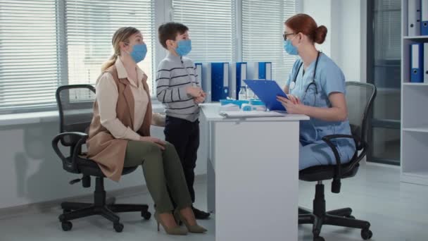 Pediatrician advises masked family at an appointment in hospital office, female physician in glasses measures the temperature of little patient — Stock video