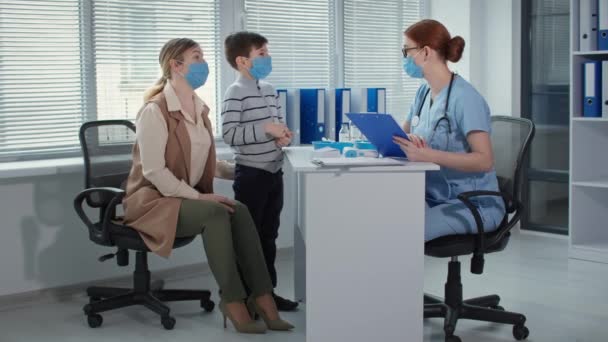 Pediatrician advises masked family at an appointment in hospital office, female physician in glasses measures the temperature of little patient — Stockvideo