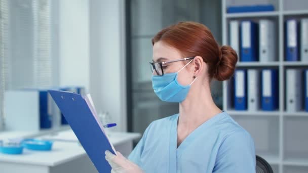 Retrato de médico en uniforme azul escribe notas en portapapeles, terapeuta en máscara protectora mirando a la cámara en el hospital — Vídeo de stock