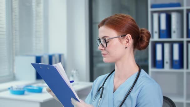 Ärztin in Schutzhandschuhen schreibt Notizen ins Klemmbrett, glückliche Therapeutin mit Brille und blauer Uniform blickt in die Kamera im Krankenhaus — Stockvideo