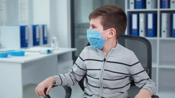 Child in mask looks at camera, portrait of a little patient sitting in a chair in health clinic on the background of many folders — Stockvideo