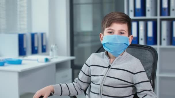 Masked little boy, portrait of patient sitting in chair at the hospital office against the background of many folders — Stok video