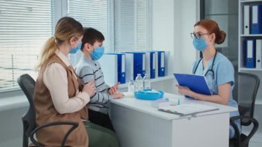 family consults the attending physician while sitting in the hospital office, medical doctor in mask and glasses writes on clipboard at table