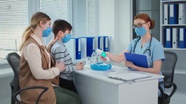 visit to doctor with mom, masked female pediatrician measures the temperature of cute little boy patient with non-contact thermometer then shows the vaccine in a glass ampoule