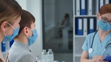 pediatrician in hospital, masked female doctor in glasses measures the temperature of boy patient with non-contact thermometer