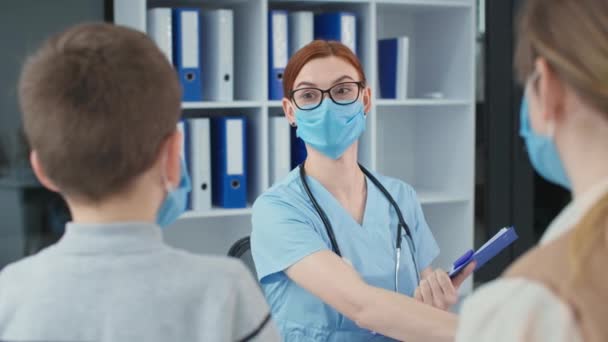 Médico en máscara protectora sentado en una mesa en el hospital, mujer joven en gafas escribe en el portapapeles y mide la temperatura del niño con un termómetro sin contacto — Vídeo de stock