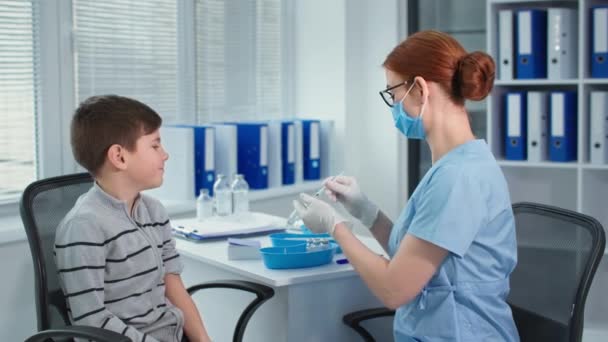 Вакцинація, лікар в захисних рукавичках і окулярах, що друкують вакцину шприцом, потім ін'єкції пацієнта хлопчика в лікарняному кабінеті під час карантину — стокове відео