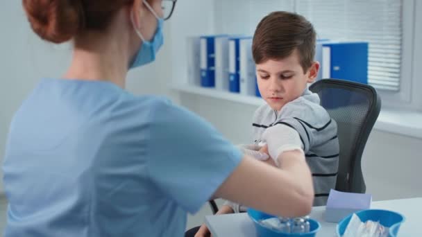 Childhood vaccination, masked doctor in protective gloves giving an injection to little patient sitting in a chair at hospital office — Stok video