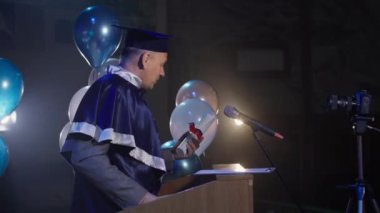 Akademik şapka takan çekici erkek öğretmen, meclis salonunda kürsünün arkasında dururken mezuniyet törenini yönetiyor.