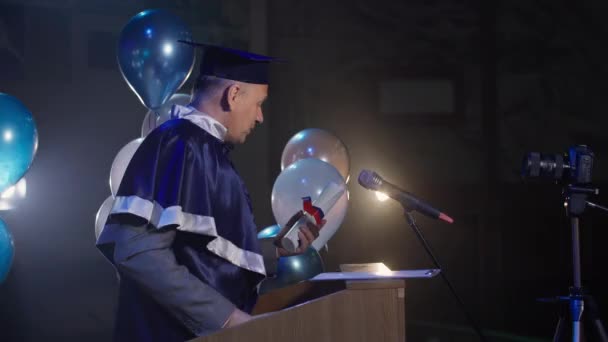 Atraktivní mužský učitel v akademické čepici a plášti provádí promoční ceremoniál, zatímco stojí za řečnickým pultem v montážní hale — Stock video