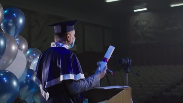 Professeur d'université en vêtements académiques et chapeau en masque médical présente des diplômes à l'aide de caméra vidéo dans la salle de conférence — Video
