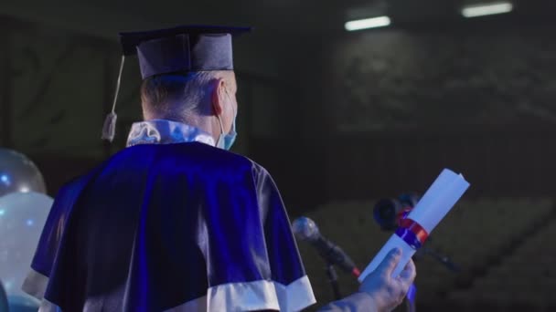 Professor do sexo masculino r em manto acadêmico e chapéu em máscara médica apresenta diplomas para graduados perto de microfone via link de vídeo na sala de conferências — Vídeo de Stock