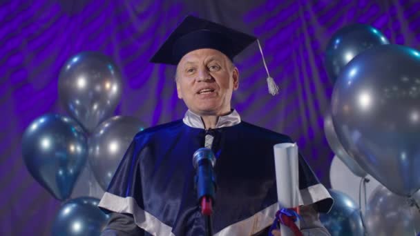 O homem na roupa acadêmica com o diploma em mãos conduz a cerimônia de graduação cerimonial e congratula graduados no contexto de microfone de balões no corredor de reunião, olha para a câmera — Vídeo de Stock