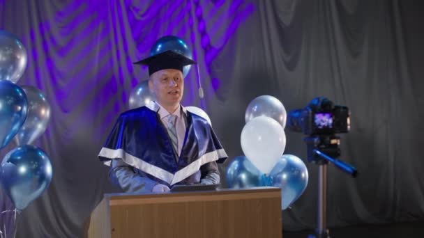 Volwassen mannelijke rector van het instituut voert ceremoniële deel van de presentatie van het diploma aan studenten met behulp van video link als gevolg van quarantaine en pandemie achtergrond van ballonnen — Stockvideo