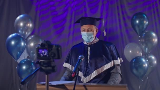 Преподаватель-мужчина в медицинской маске соблюдает меры предосторожности и поздравляет выпускников с окончанием учебы онлайн по видеосвязи во время пандемии и карантина — стоковое видео