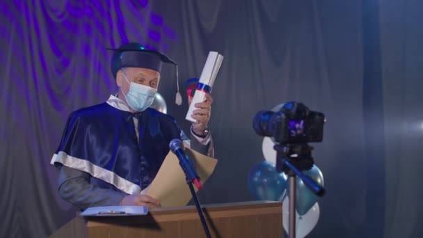 Профессор в медицинской маске проводит церемонию вручения дипломов онлайн по видеосвязи в конференц-зале, пандемия — стоковое видео