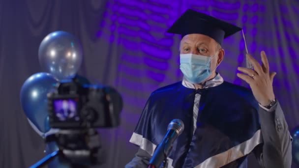Männlicher Dozent in Mantel und medizinischer Maske gratuliert Absolventen feierlich zum Ende des akademischen Jahres bei der Online-Abschlussfeier im Konferenzraum stehend und beobachtet moderne — Stockvideo