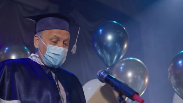 Gecelik ve tıp maskesi takan üniversite erkek profesörü, konferans salonunda düzenlenen tören sırasında öğrencilere resmi olarak diplomaları sunuyor. — Stok video