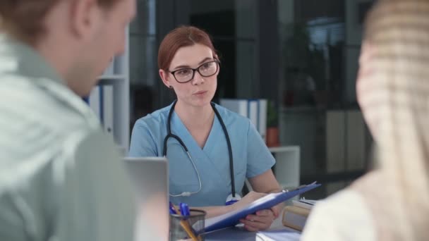 Orvos orvosi ruhák fonendoszkóppal ül asztalhoz, és konzultál férfi és nő egészségi állapotáról klinikákon iroda — Stock videók