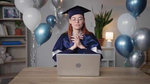 Joven estudiante alegre en traje académico emocionalmente se alegra de graduarse de la universidad y recibir un diploma en línea sentado en la habitación a la mesa en el fondo de globos — Vídeos de Stock