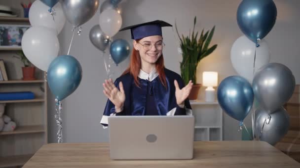 Graduación en línea, mujer joven con una gorra académica y vestido se regocija por la presentación del diploma en la ceremonia virtual a través de un enlace de vídeo en el ordenador portátil mientras se sienta en casa fondo de globos — Vídeos de Stock