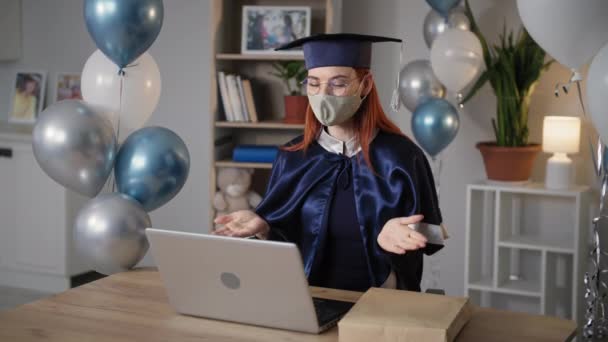 Graduation in quarantine, happy female student wearing mask on face and academic cap and gown rejoices at graduation online and gives diploma from an envelope sitting at home at laptop background of — Stock Video