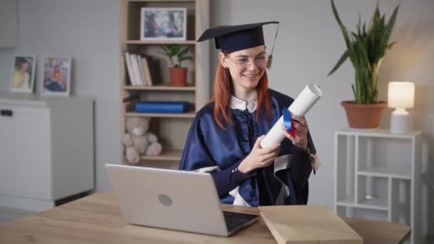 Female graduate with diploma in hands rejoices at online university graduation ceremony using webcam on laptop while sitting at home on background of air balloon — Stock Video