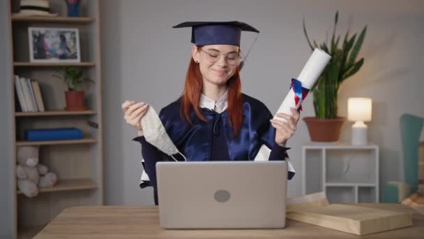 Young female student happily takes off mask from face rejoices at end of school year online sitting at laptop in room, smiling and looking at camera — Stock Video