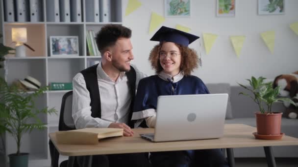現代の卒業式で魅力的な卒業生は卒業式で夫を抱きしめるラップトップのビデオリンク経由で卒業証書を受け取る — ストック動画