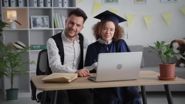 Diploma on-line, mulher jovem feliz no manto acadêmico e chapéu abraça seu namorado durante a cerimônia de formatura receber via link de vídeo no laptop — Vídeo de Stock