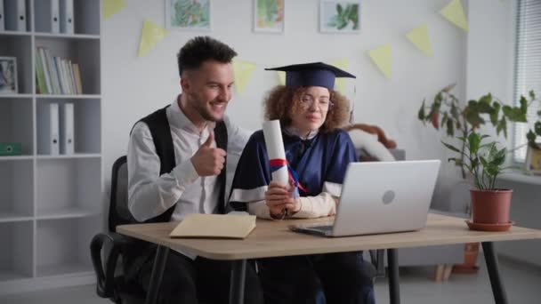 Ensino à distância, alegre jovem casal em roupas acadêmicas celebrando cerimônia de formatura e diploma por link de vídeo no laptop — Vídeo de Stock
