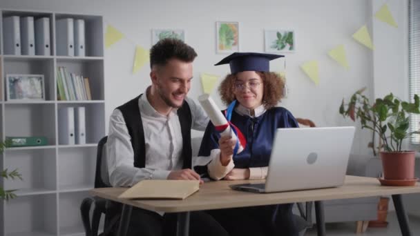 Education online, joyful girl in academic dress with friend at graduation ceremony via video link on laptop during distance learning — Stock Video